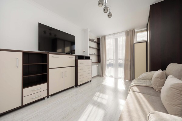 Una habitación moderna y bien iluminada con kitchenette, un televisor de pantalla plana, un sofá y un ventanal con cortinas.