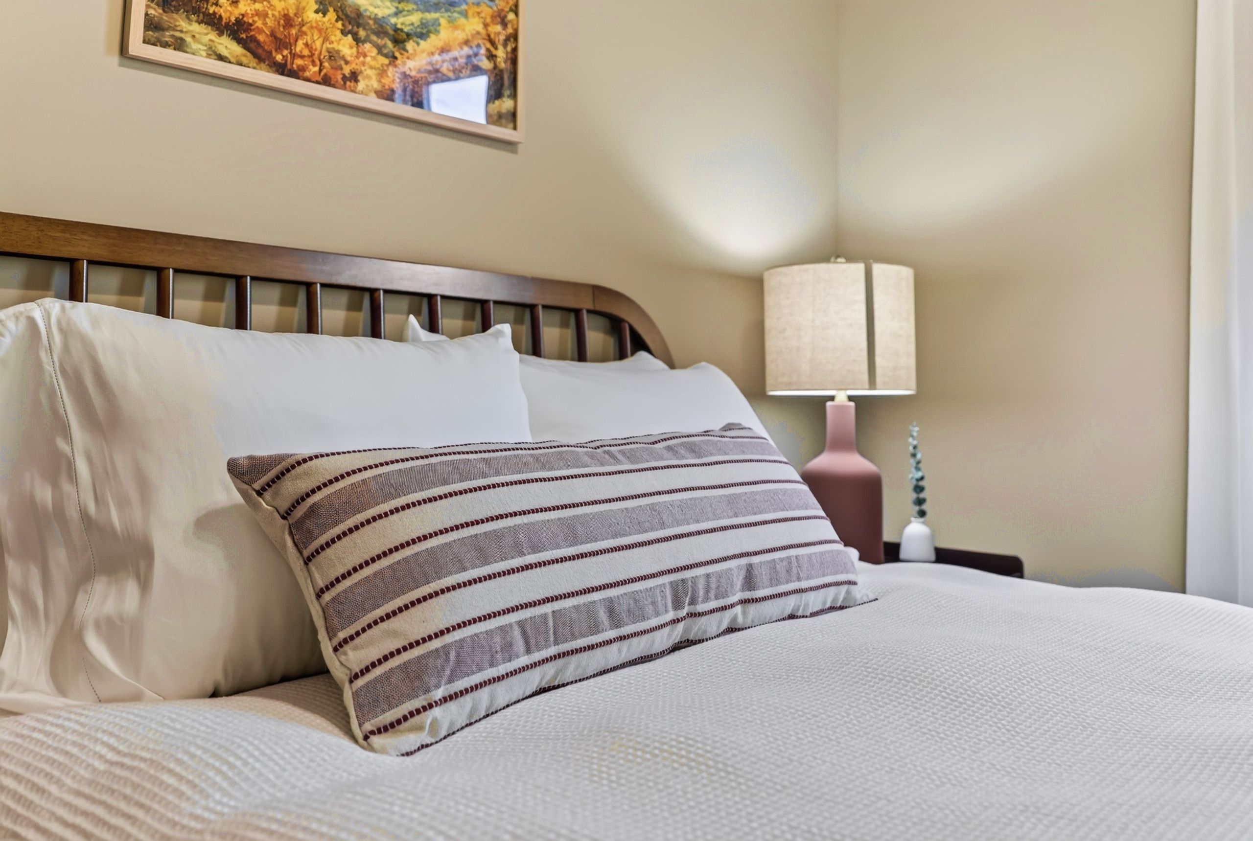 A neatly made bed with a striped pillow and a plain pillow, a wooden headboard, a wall-mounted picture, a bedside lamp, and a nightstand with a vase.