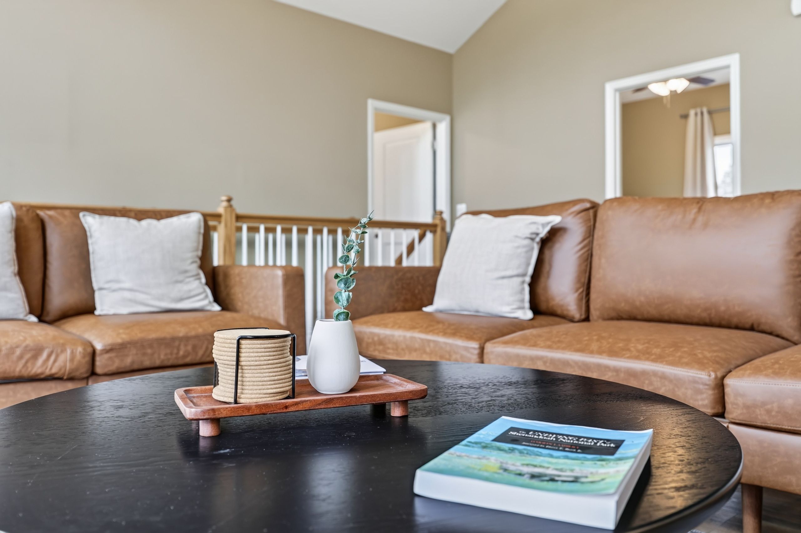 A living room with two brown leather sofas, a coffee table, a stack of books, a white vase with a plant, and a book titled 'The Great Gatsby'.