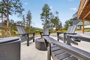 An outdoor seating area with Adirondack chairs and a fire pit, surrounded by trees and a view of a house.
