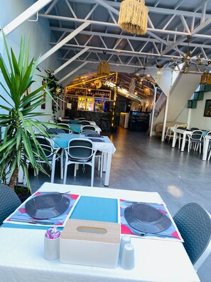 Un restaurante con techo alto y vigas a la vista, mesas dispuestas y una zona de bar al fondo.