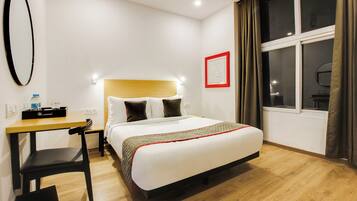 Habitación de hotel con cama, escritorio, silla, ventana con cortinas y espejo adosado a la pared.