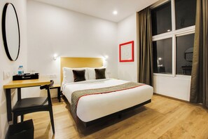 A hotel room with a bed, a desk, a chair, a window with curtains, and a wall-mounted mirror.