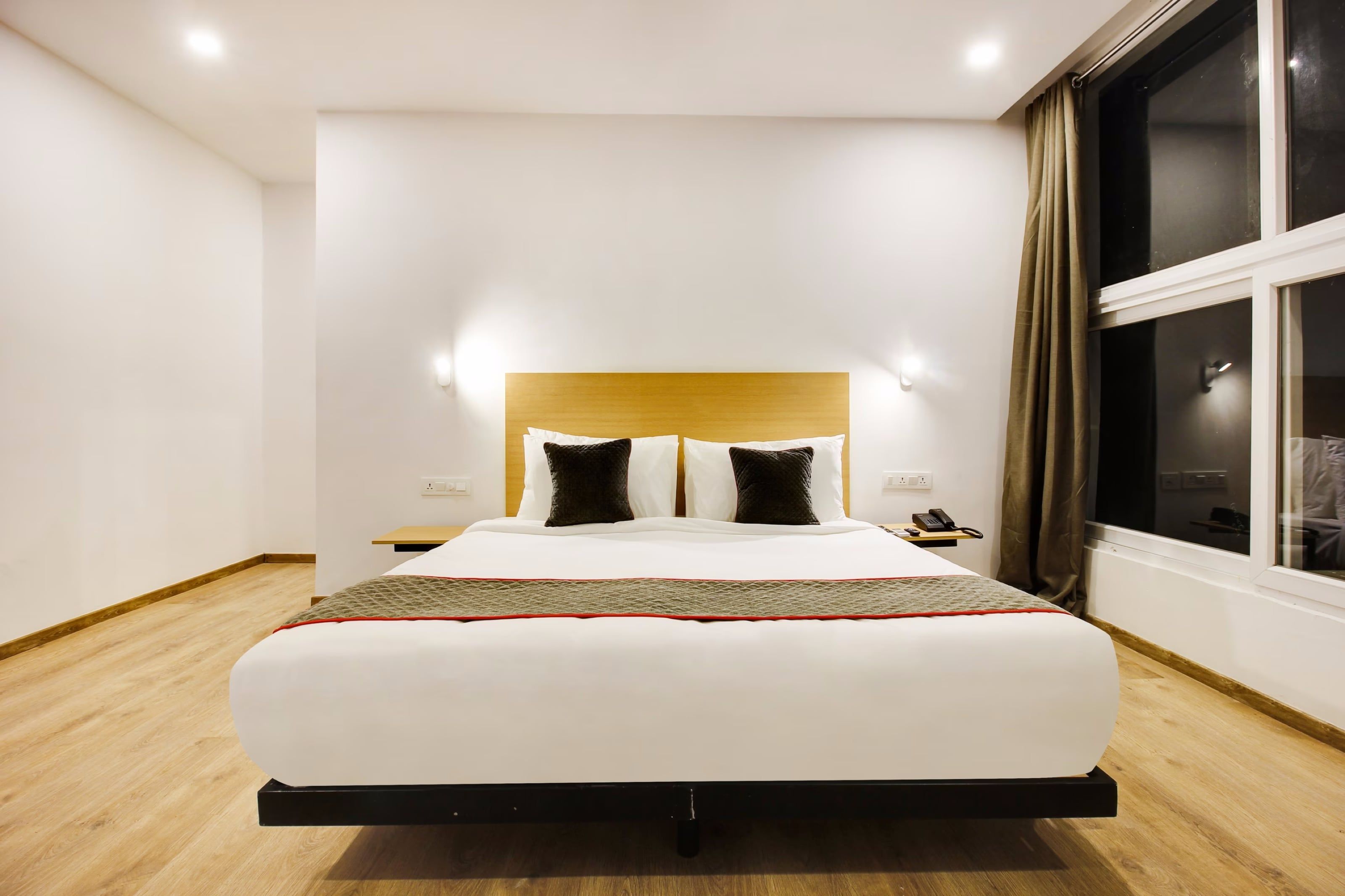 A modern hotel room with a large bed, a wooden headboard, a bedside table with a phone, and a window with curtains.