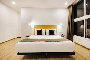 Una habitación de hotel moderna con una cama grande, cabecera de madera, mesita de noche con teléfono y ventana con cortinas.