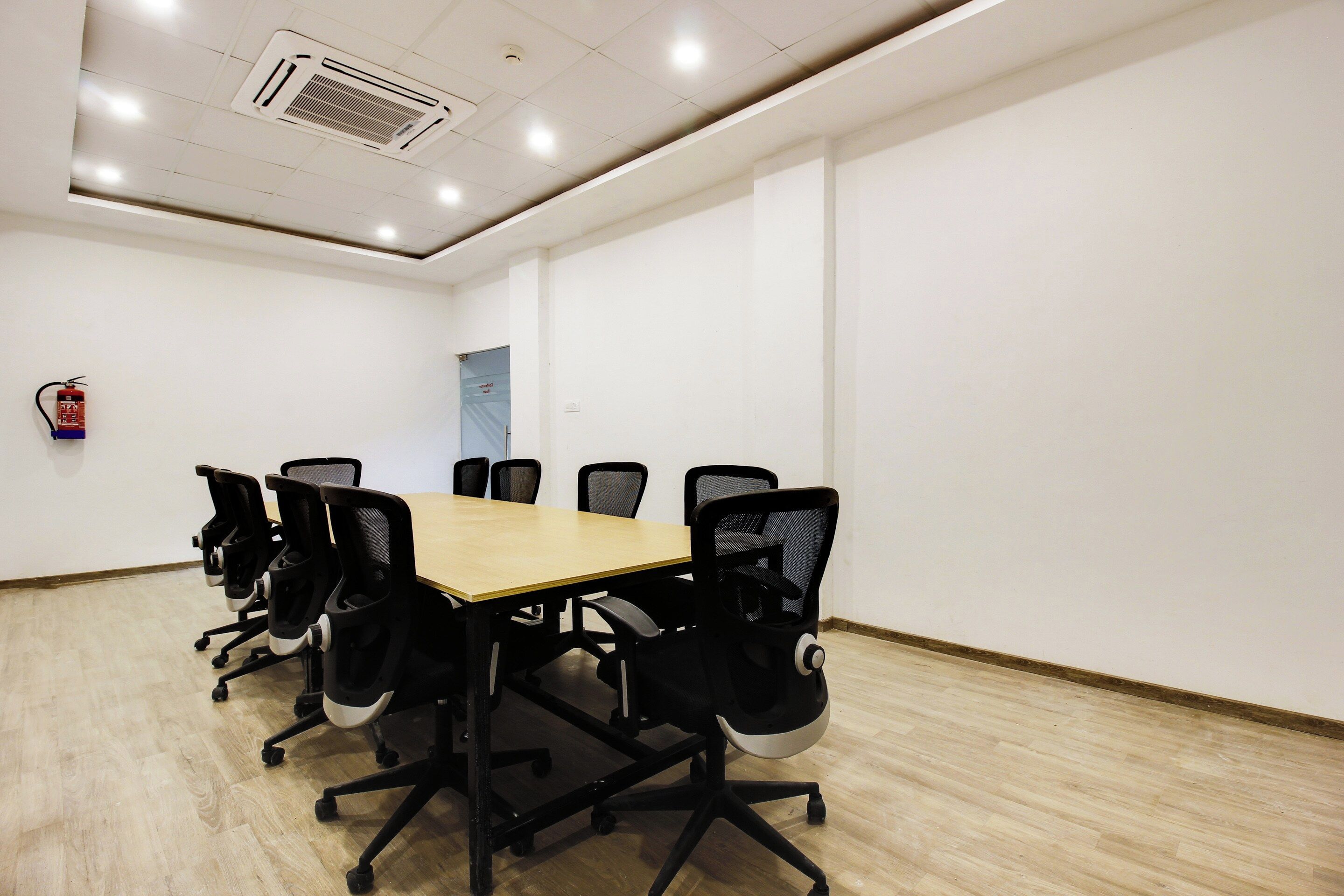 A conference room with a long table and six chairs, a fire extinguisher on the wall, and a door in the background.