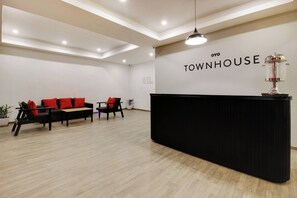 Zona de recepción con mostrador negro, área de asientos con sillas negras y cojines rojos, y un cartel en la pared que dice 'OYO TOWNHOUSE'.