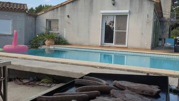 Une piscine avec un barbecue au premier plan, un bâtiment avec un toit en tuiles et une porte avec un panneau de verre.