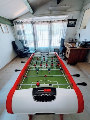 Una habitació amb taula de futbolí, escriptori amb ordinador i finestra amb cortines.