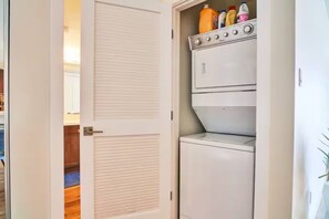 Ein Waschraum mit einer weißen Waschmaschine und einem Trockner, einem Schrank für Reinigungsmittel und einer Tür zu einem anderen Raum.