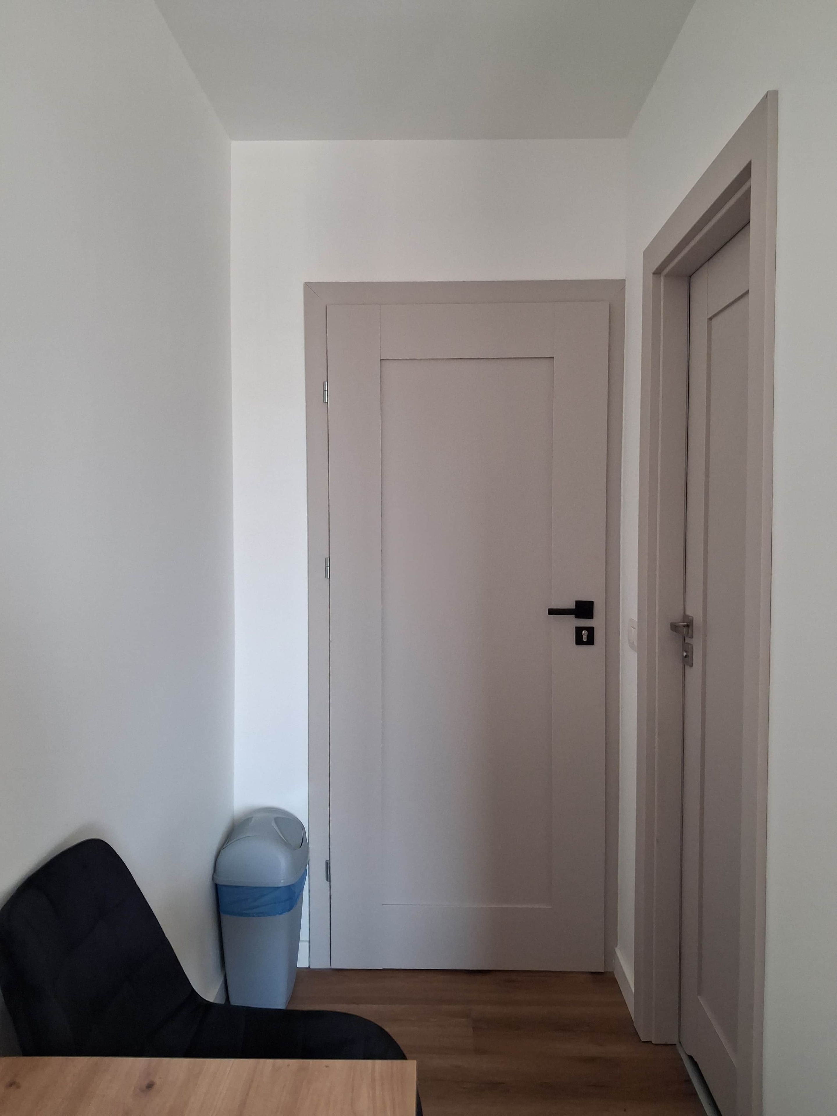 Un couloir avec une porte fermée, une poubelle et une chaise.