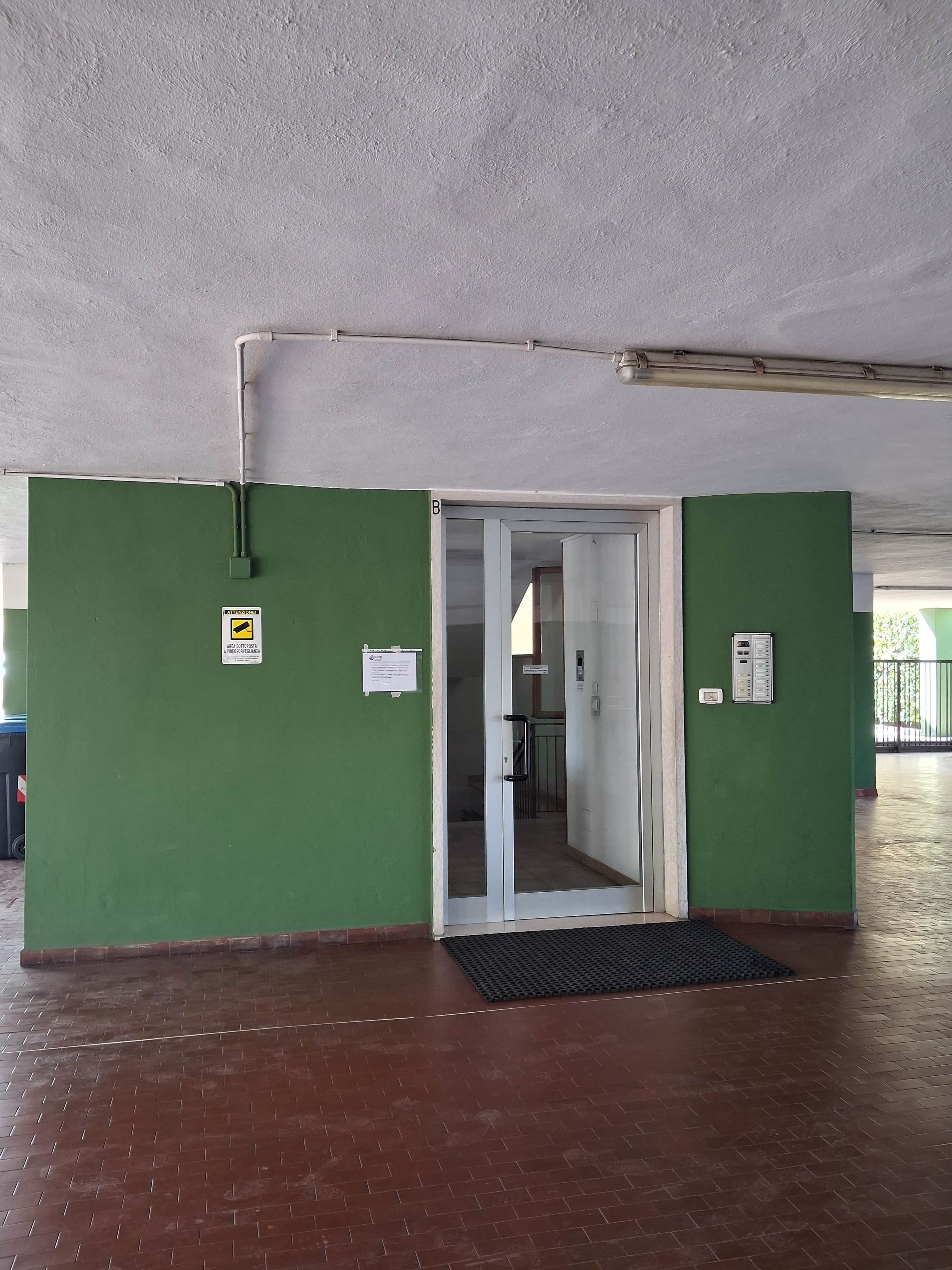 A hallway with green walls, a tiled floor, and a door leading to another room. There are signs on the walls and a vent on the ceiling.