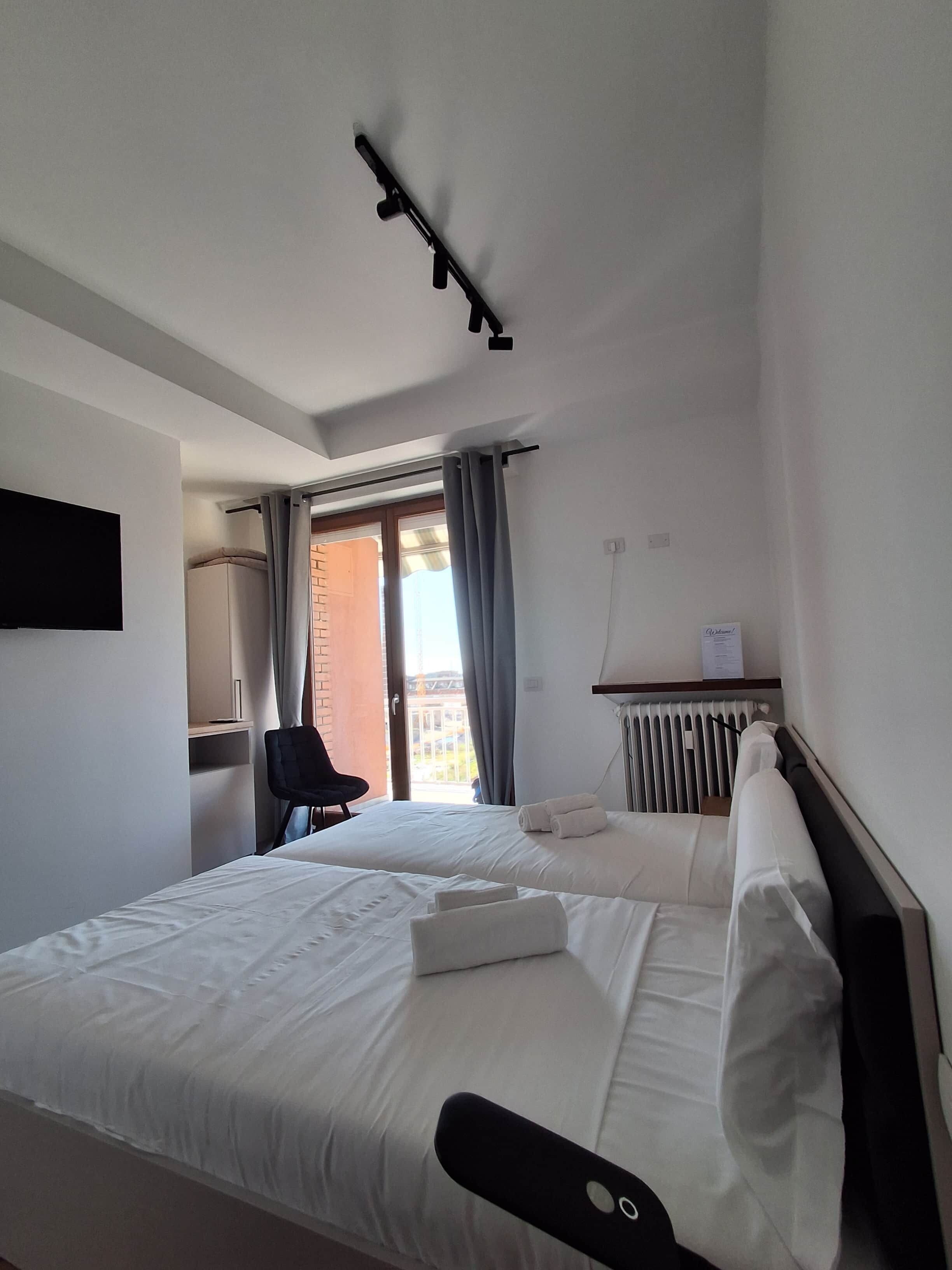 A hotel room with a bed, a chair, a desk, a radiator, and a window with curtains.
