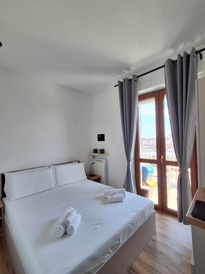 A hotel room with a bed, a nightstand, a lamp, a window with curtains, and a view of the outside. - Porta Fiera | Room 3 (Verona)