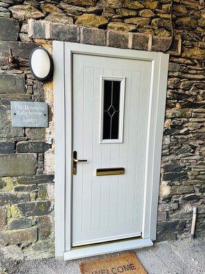 Eine weiße Tür mit einem schwarzen Glasfeld und einem Metallgriff, vor einer Steinwand, auf der ein Schild mit der Aufschrift 'The Bowness Lakehouse Lodge' steht.