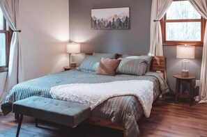 A bedroom with a large bed, two bedside lamps, a window with curtains, and a painting on the wall. - Hot Tub, Game Room, Fire Pit Cabin, Sleeps 10 near Old Man's Cave (Logan)
