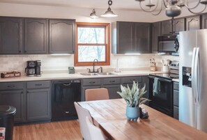 A modern kitchen with dark cabinets, a wooden dining table, a coffee bar, and a window with a view of bare trees. - Bigfoot's Hideout - Cabin-Game Room Sleeps 10 (Logan)