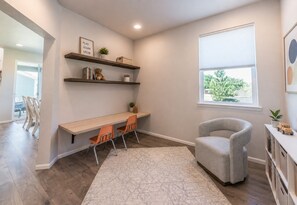 Une pièce comprenant un bureau, deux chaises, un fauteuil gris, une fenêtre avec des stores et des étagères remplies de livres et d’un ours en peluche.