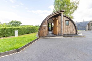 A wooden structure with a door and a small window, surrounded by a paved area and greenery. - Charming Stay In A Mid Wales River Escape (Llangammarch Wells)