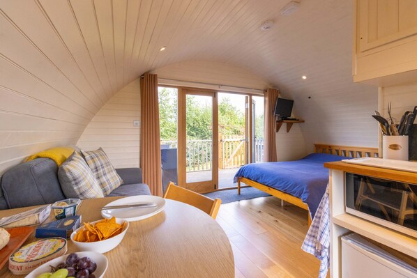 A cozy room with a bed, a sofa, a dining table with snacks, and a TV. - Cosy Glamping Pod Near Garth With River Views (Llangammarch Wells)