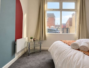 Une chambre à coucher avec un lit, une petite table sur laquelle est posée une plante, un radiateur et une fenêtre avec des rideaux.