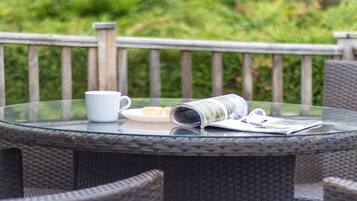A round glass table with wicker chairs, a white mug, a plate with pastries, and a rolled-up magazine.