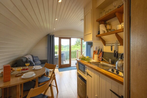 A compact kitchen with a dining area, a built-in oven, and a view of the outdoors. - Riverside Glamping Pod With Free Wifi Parking (Llangammarch Wells)