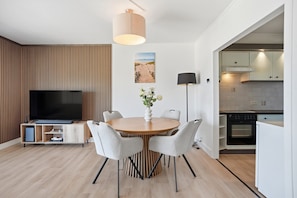 Ein modernes Wohnzimmer mit einem Flachbildfernseher, einem runden Esstisch mit Stühlen, einer Stehlampe und einem Gemälde mit Strandmotiv.