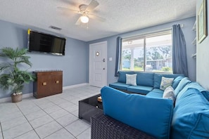 A living room with a blue sofa, a television mounted on the wall, a ceiling fan, and a window with curtains. - Oyster Bay Escape | Private Dock, Ramp & Kayak (New Port Richey)