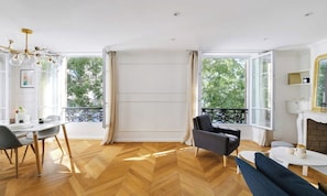 Een ruime woonkamer met een houten parketvloer, een eethoek met een witte tafel en stoelen, een zwarte fauteuil en een bank. Grote ramen bieden uitzicht op groen buiten.