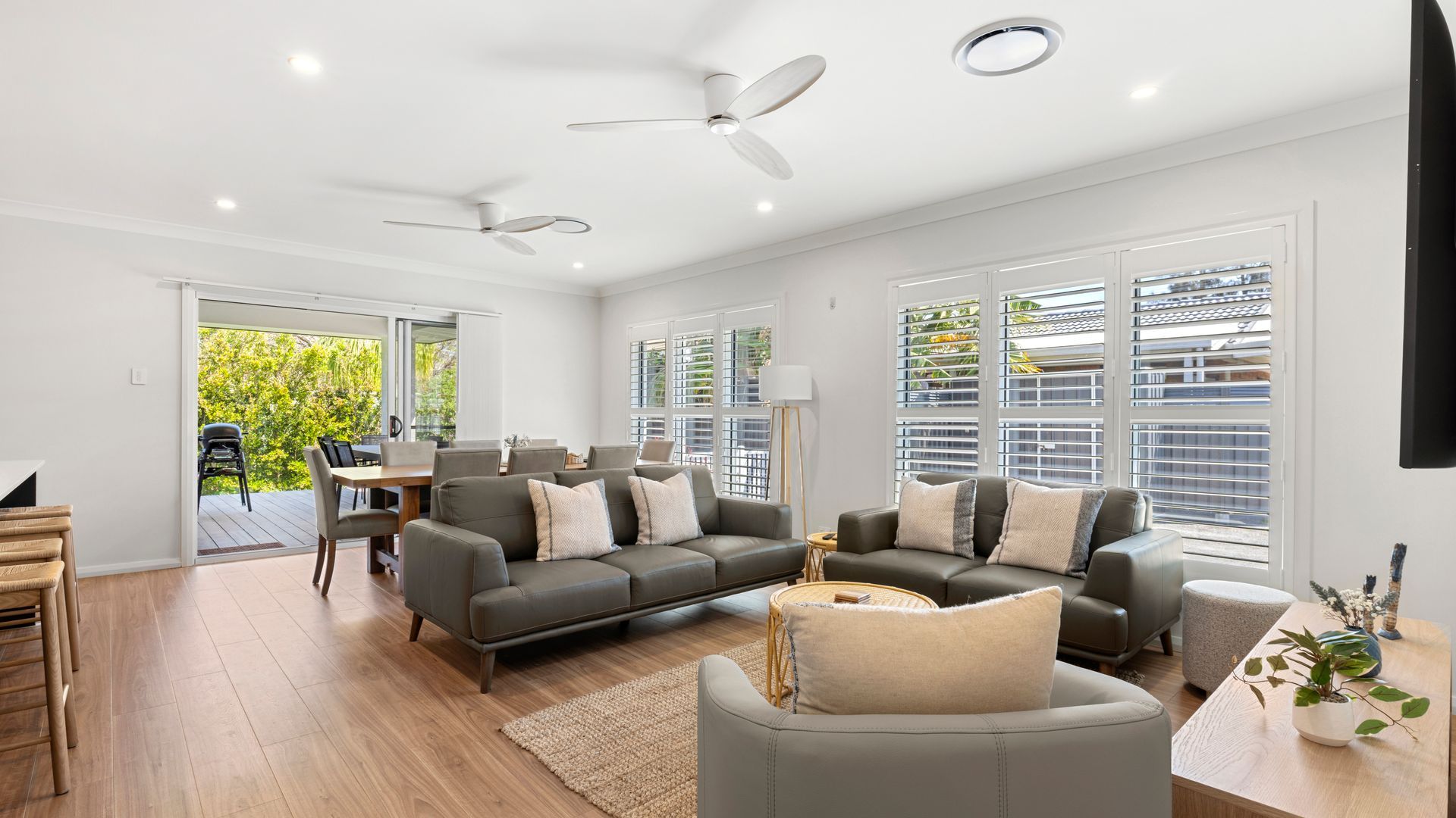 A spacious living room with a grey sofa set, a dining area with a table and chairs, and large windows with blinds.