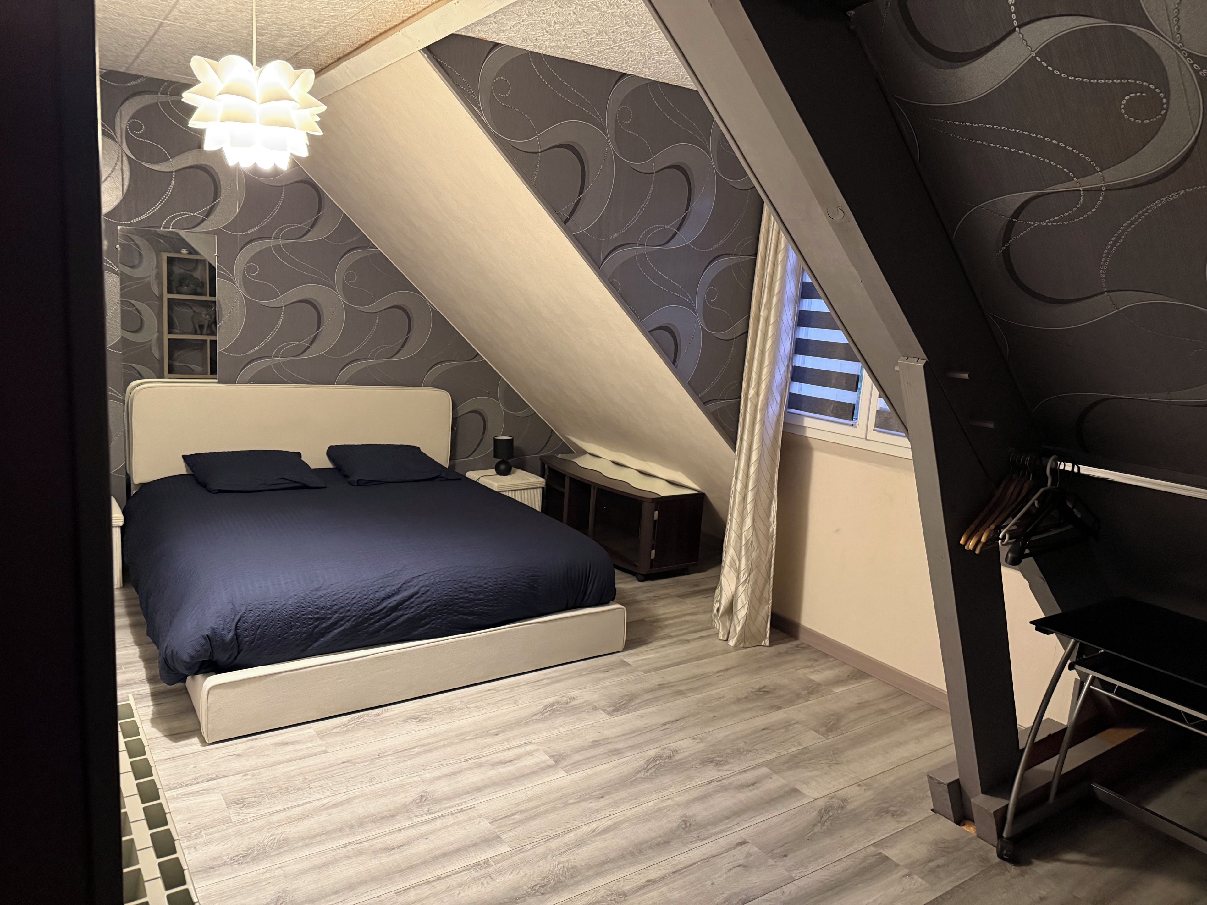 Une chambre à coucher avec un lit, une table de chevet, un luminaire suspendu et une fenêtre avec des rideaux.
