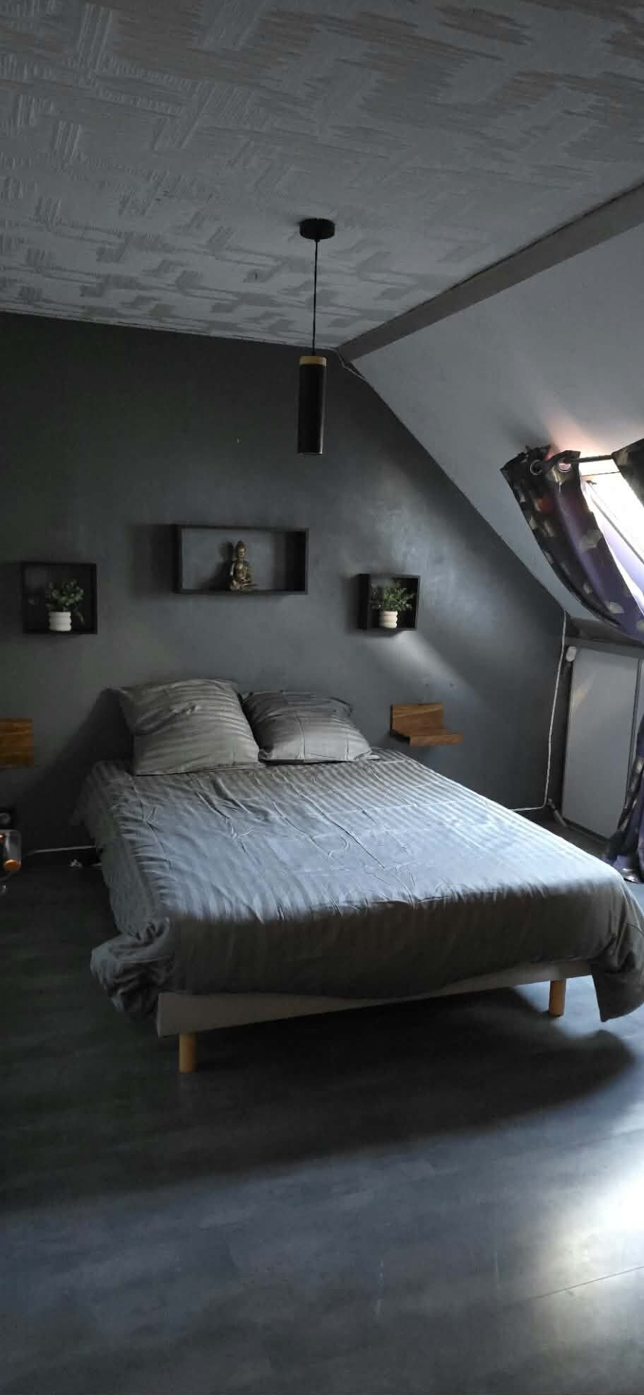 A bedroom with a sloped ceiling, a bed with a grey bedspread, two bedside tables with lamps, and wall-mounted shelves with decorative items.