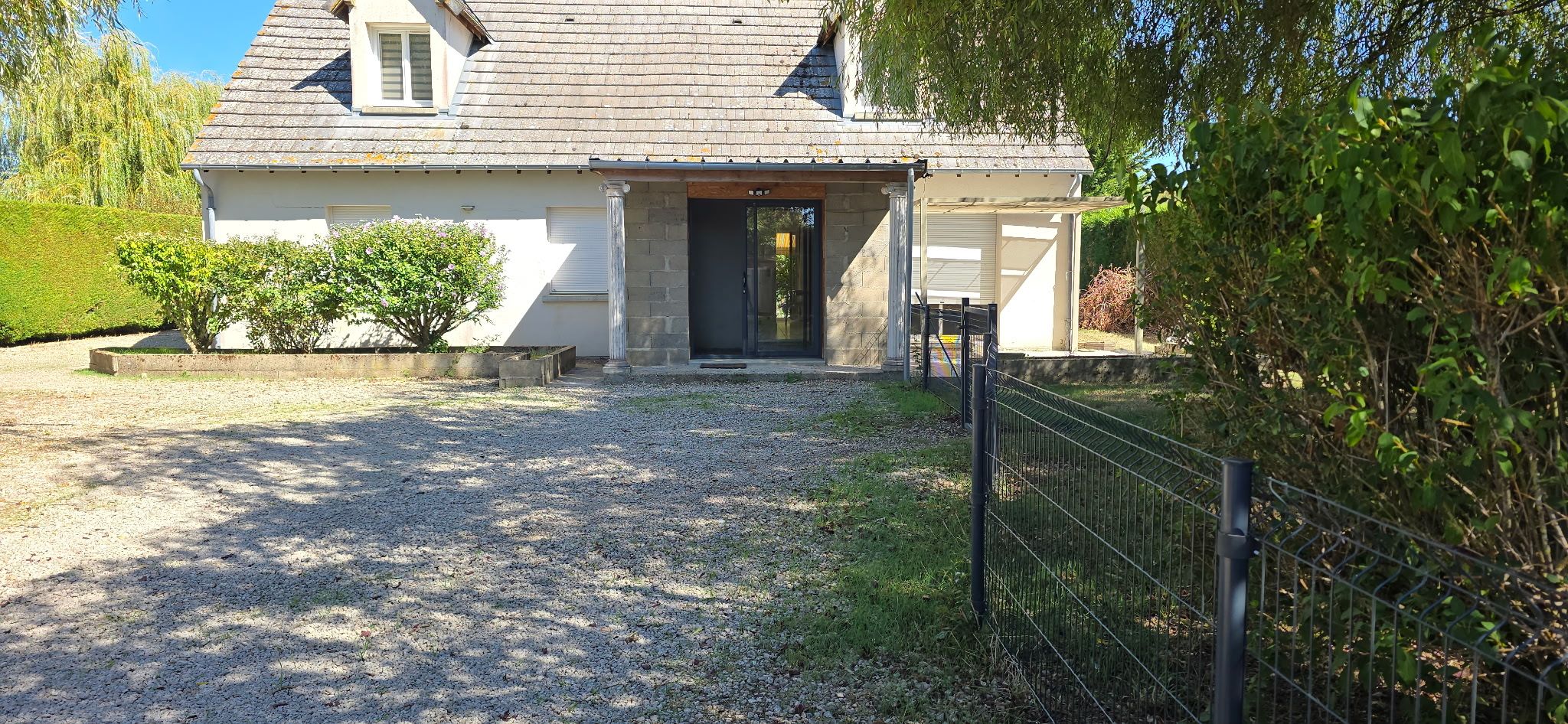 Una casa con camino de grava que lleva a una puerta de entrada oscura, rodeada de follaje y una valla metálica.