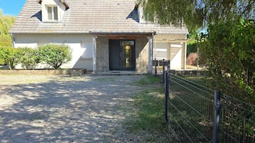Una casa con camino de grava que lleva a una puerta de entrada oscura, rodeada de follaje y una valla metálica.