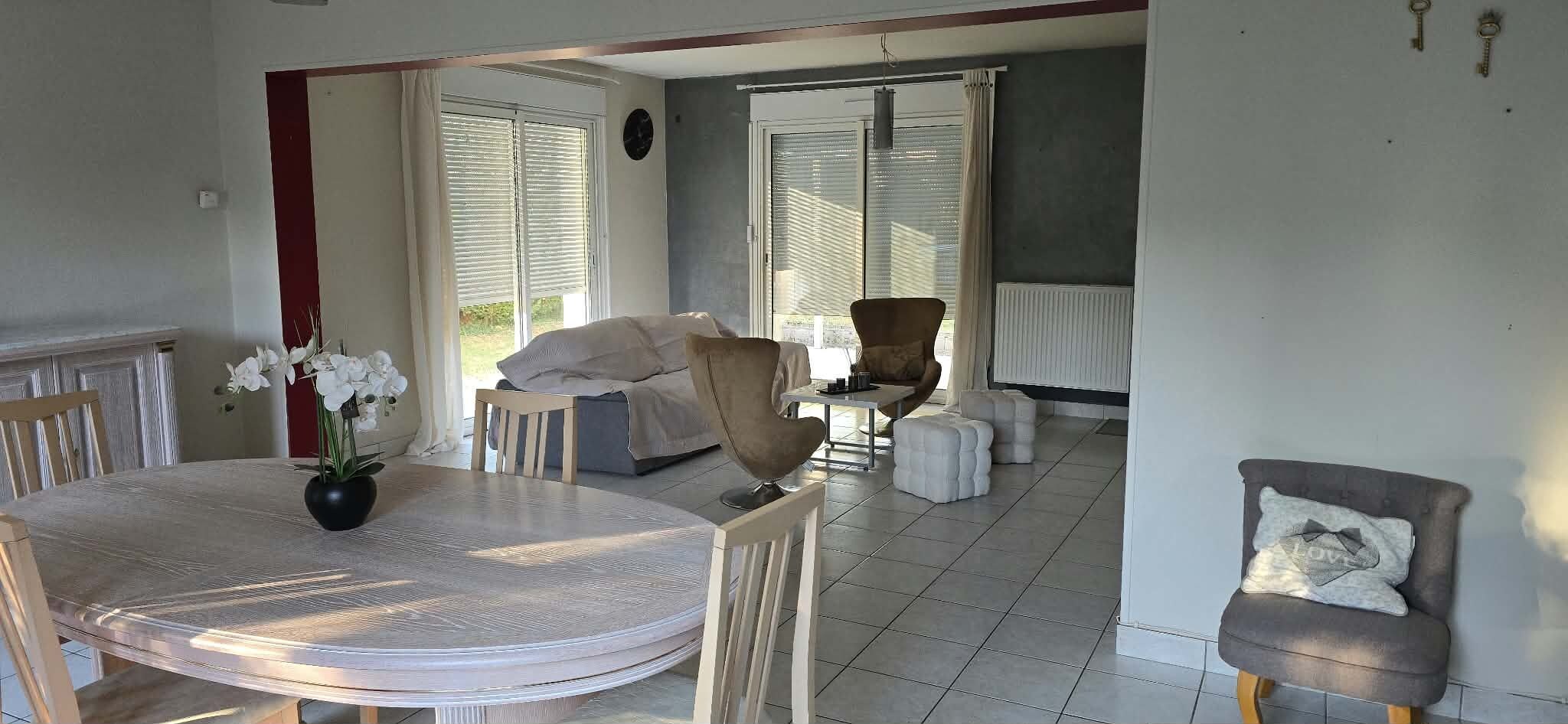 A dining area with a round table, chairs, and a potted plant. There is a sofa and a chair in the background.
