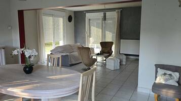 A dining area with a round table, chairs, and a potted plant. There is a sofa and a chair in the background.