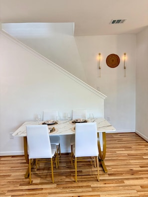 Uma área de refeições com uma mesa branca posta para quatro pessoas, piso de madeira e uma escada branca.