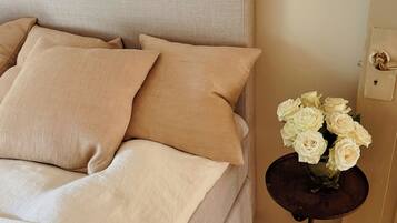 Een bed met beige beddengoed en kussens, een nachtkastje met een vaas witte bloemen, en een wandlamp.