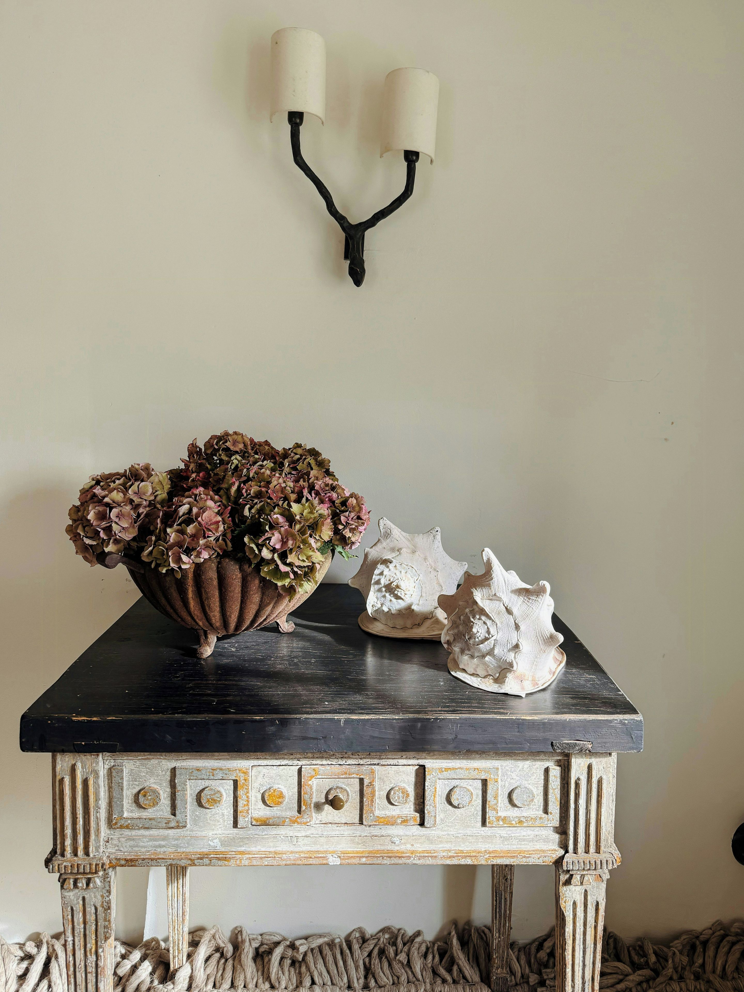 A table with a decorative bowl of flowers, a shell, and a wall-mounted light fixture.