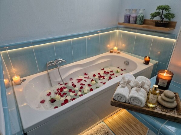 A bathroom with a bathtub filled with rose petals, candles, and a tray with towels and a bottle.