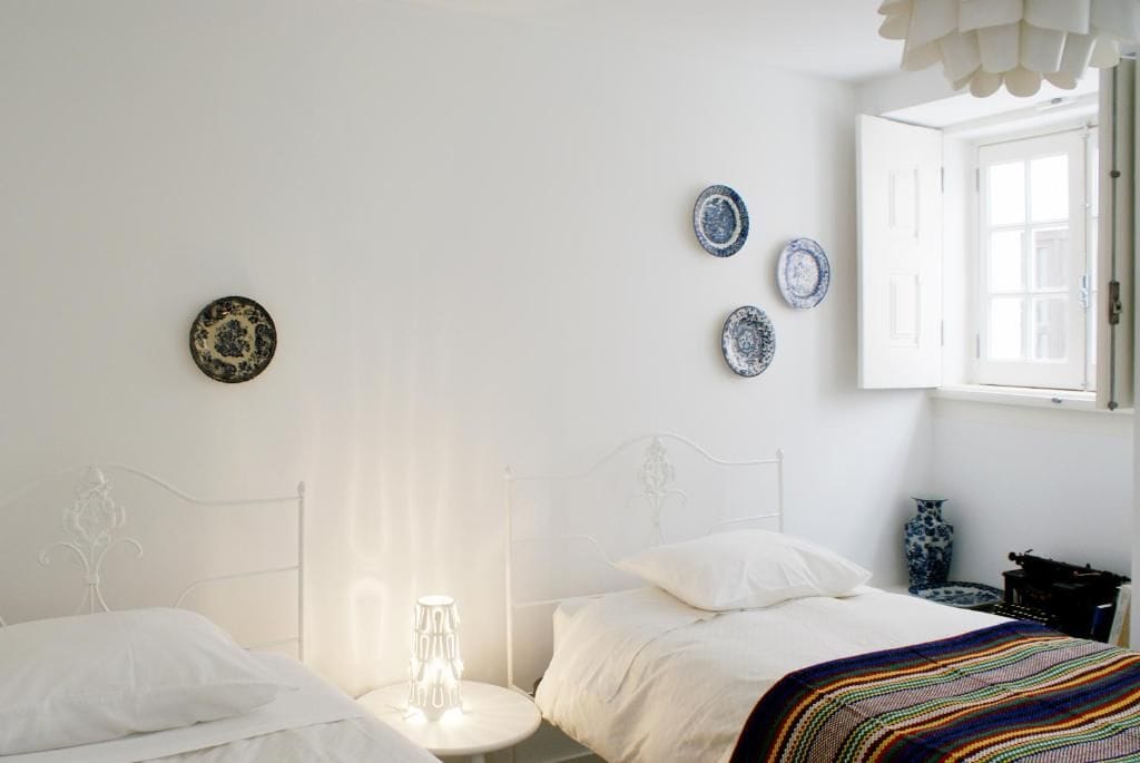 Un dormitorio con estructura de cama metálica blanca, dos almohadas blancas, una manta colorida a rayas, una mesita de noche con lámpara y tres platos decorativos en la pared.