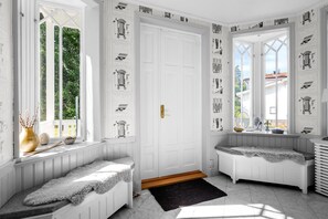 Ein Zimmer mit einer weißen Tür, zwei Fenstern mit weißen Rahmen, einer Bank mit Teppich und einer Wand mit architektonischen Zeichnungen.