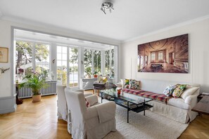 Ein geräumiges Wohnzimmer mit einer Couch, Sesseln, einem Couchtisch und einem großen Fenster mit Blick auf Bäume.