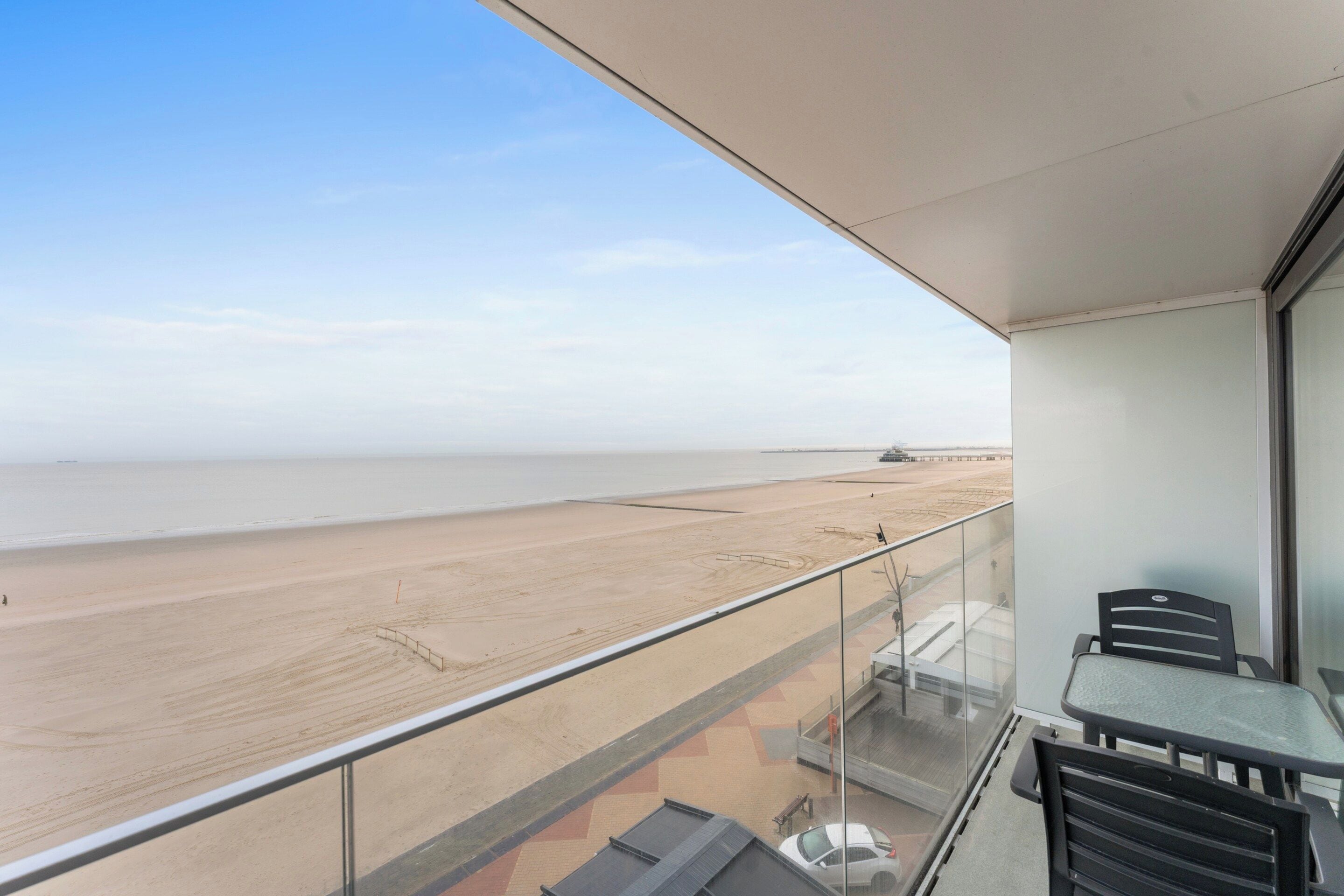 A balcony with a view of a beach and the sea. There are chairs and a table on the balcony.