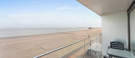 A balcony with a view of a beach and the sea. There are chairs and a table on the balcony.