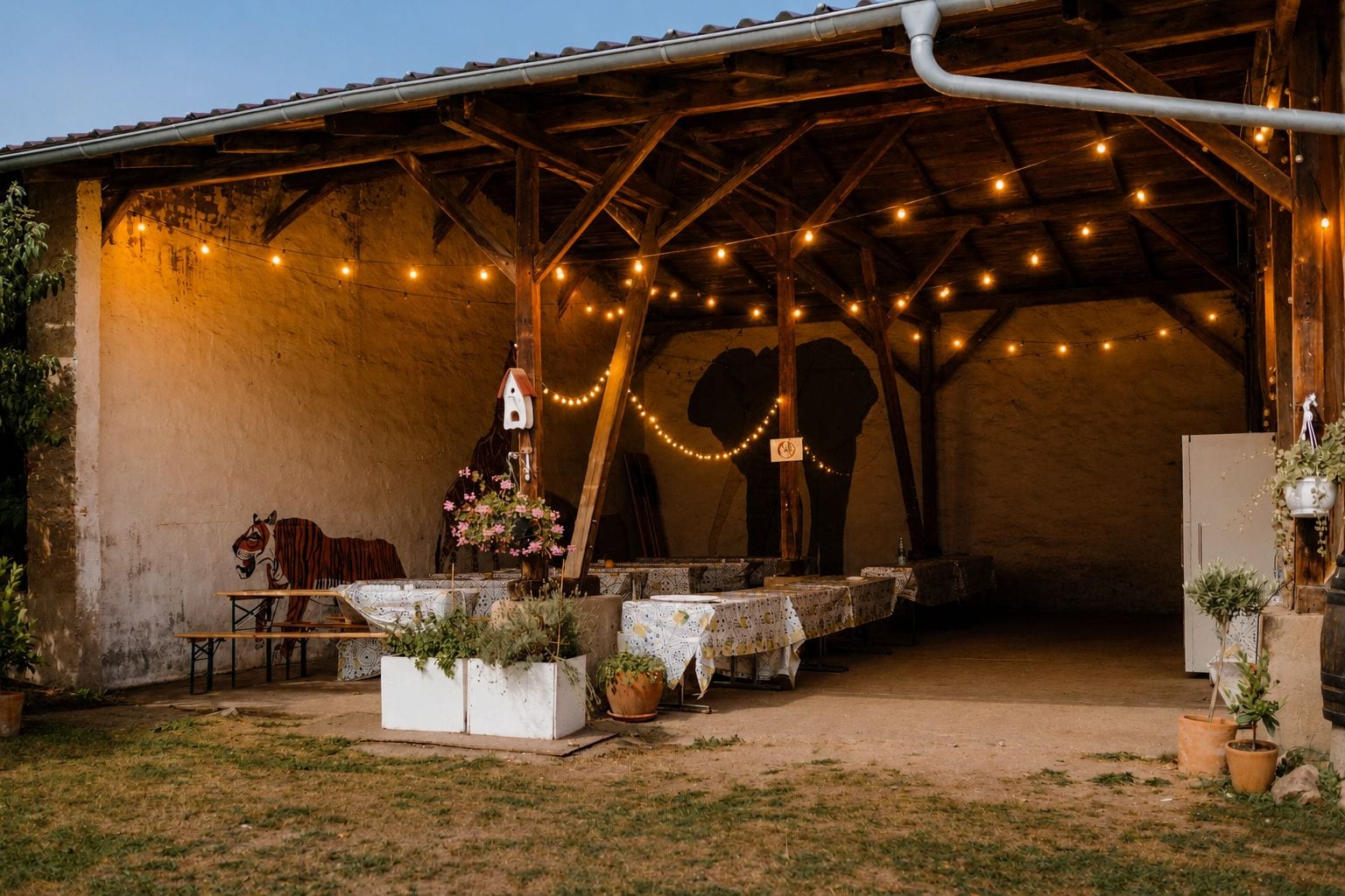 Un área de comedor al aire libre con mesas, luces colgantes y plantas en macetas.