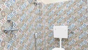 Casa de banho com painel de azulejos estampados, sanita branca, chuveiro com chuveiro manual e termoacumulador fixado na parede.