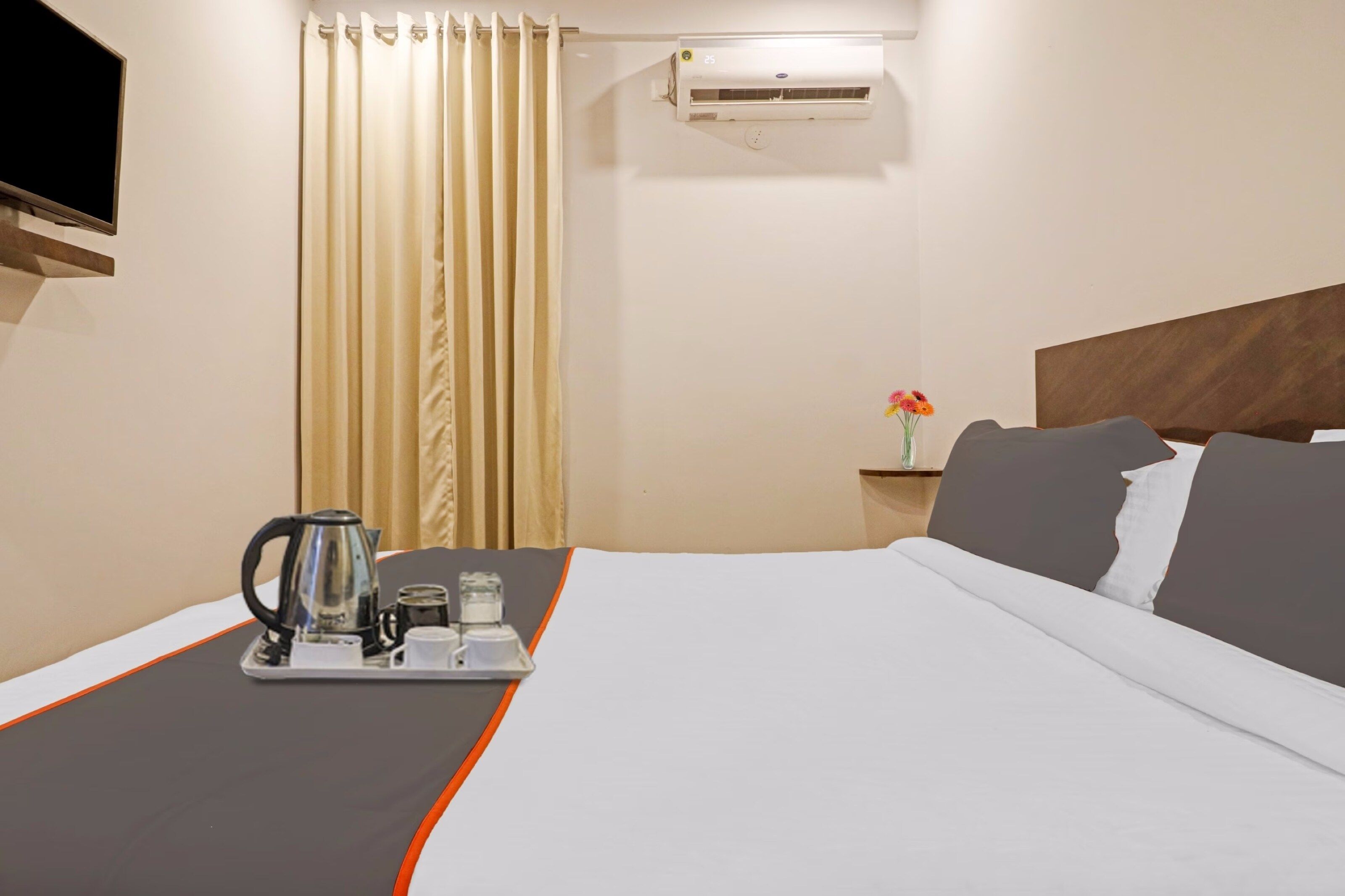 Quarto de hotel com uma cama, uma mesinha de cabeceira com um vaso de flores, um ar condicionado e um tabuleiro com chaleira e chávenas.