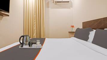 Quarto de hotel com uma cama, uma mesinha de cabeceira com um vaso de flores, um ar condicionado e um tabuleiro com chaleira e chávenas.